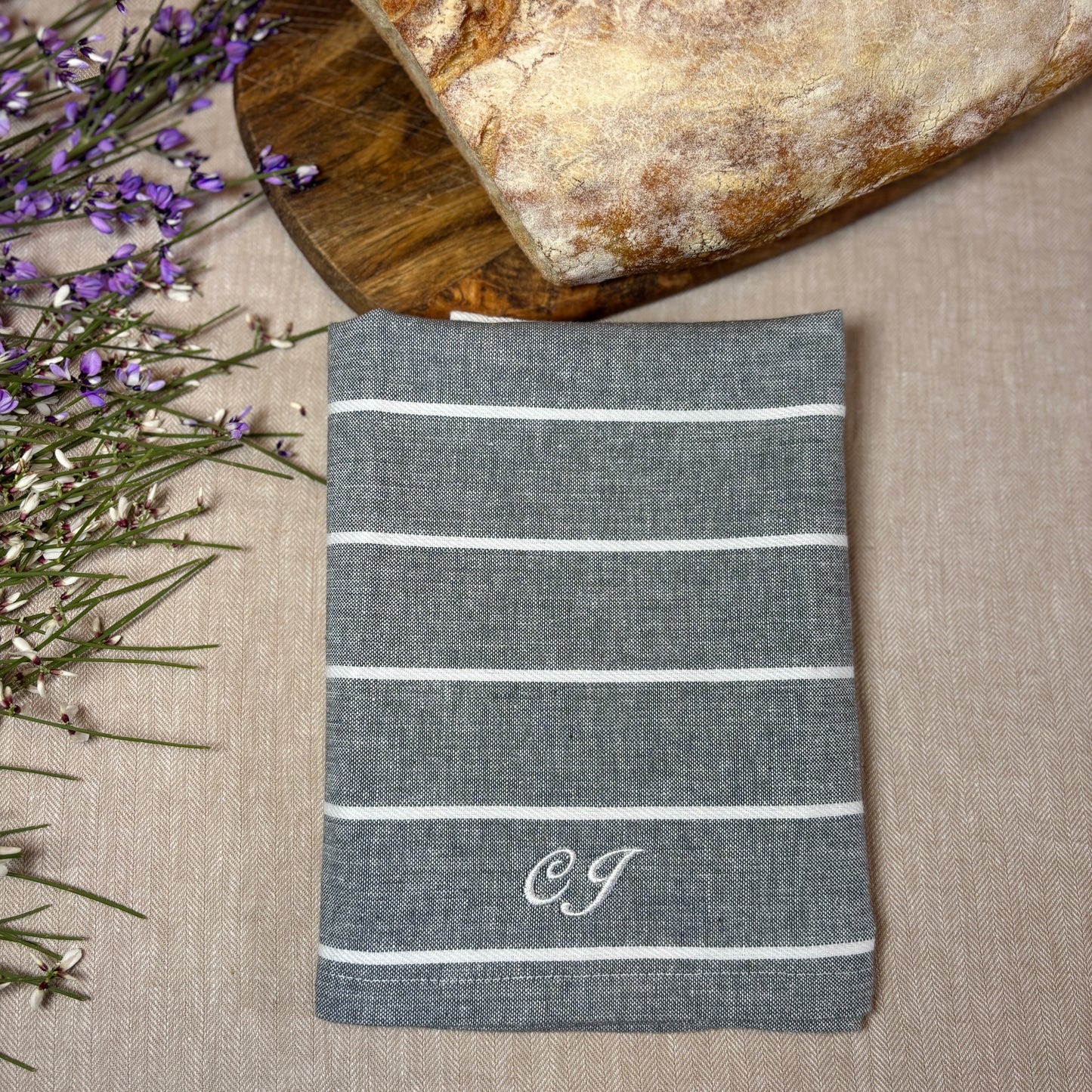 Grå tygservett med smala vita ränder och broderade initialer ligger på en beige duk med nybakat bröd och några blommor. Broderat initialer med vit brodyrtråd, CJ.