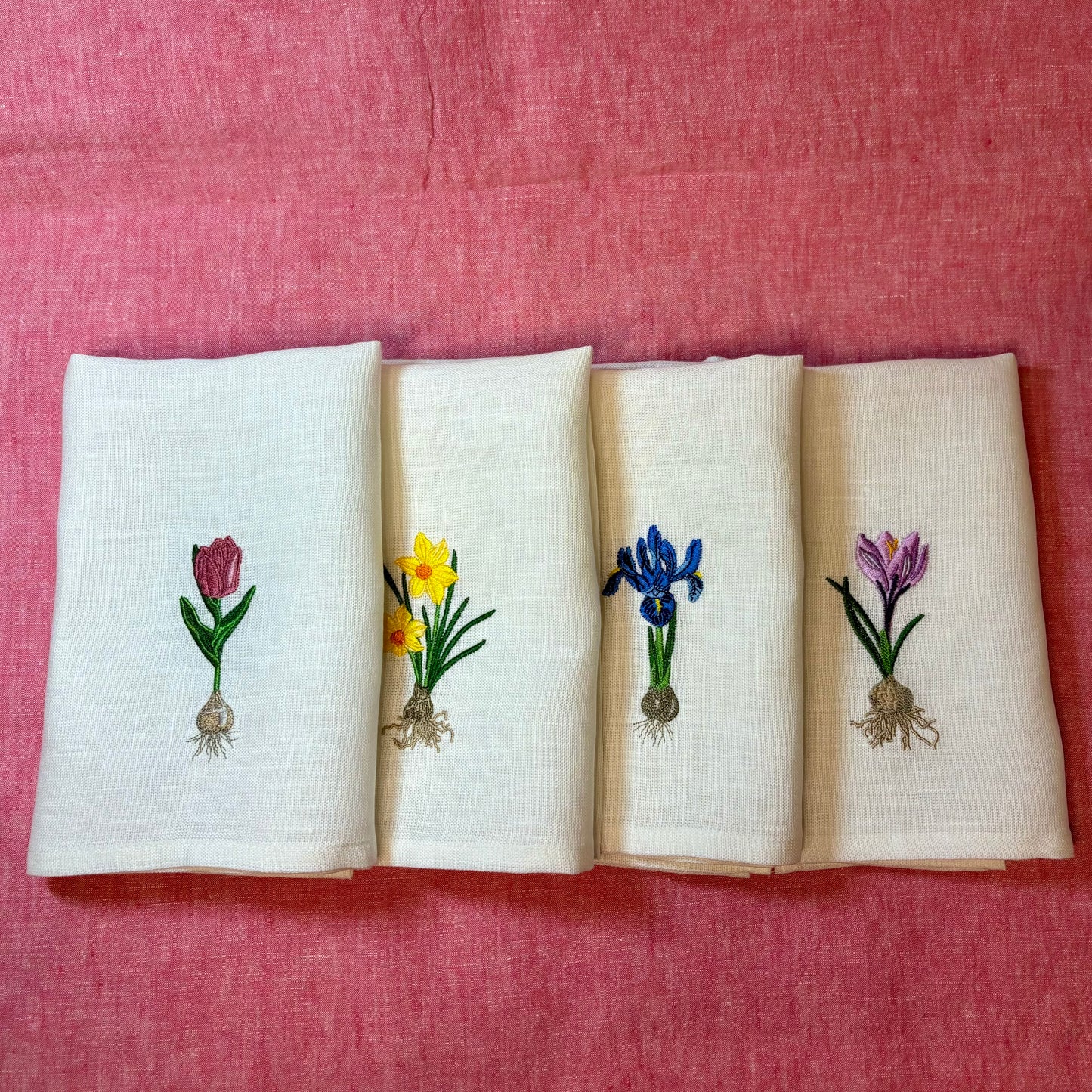 Linneservetter med fyra olika vårblommor vårlökar broderade. Blå iris, gul påsklilja, rosa tulpan och lila krokus. Alla blommor har sina lökar med. Ett set tygservetter med vårtema. Ligger på rosa linneduk