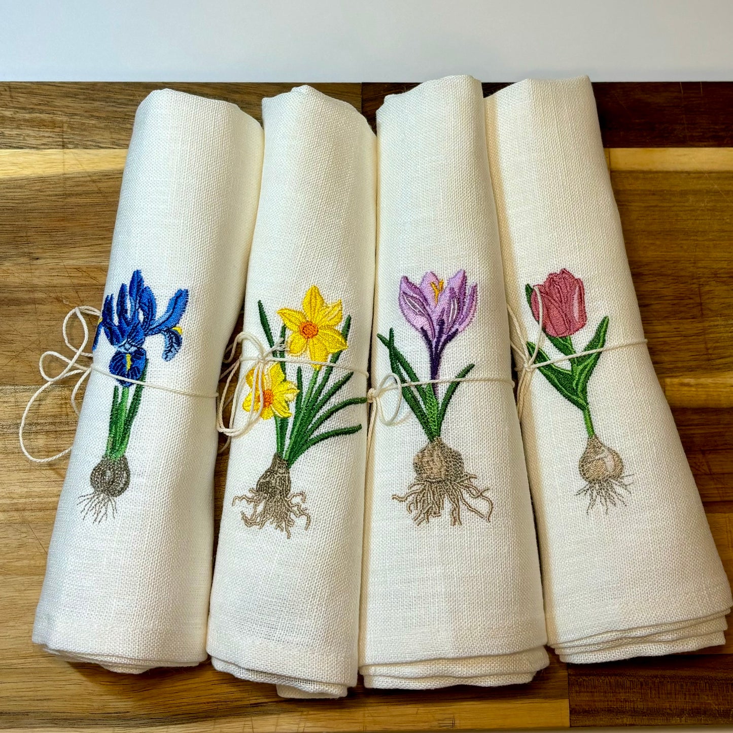 Linneservetter med fyra olika vårblommor vårlökar broderade. Blå iris, gul påsklilja, rosa tulpan och lila krokus. Alla blommor har sina lökar med. Ett set tygservetter med vårtema. Servetterna är placerade på en skärbräda av trä.