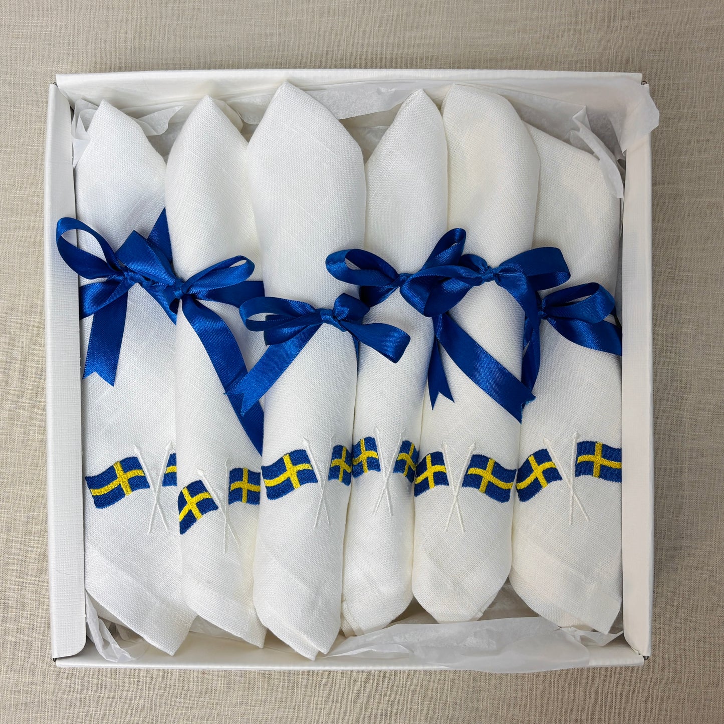 Krämvit presentask är öppen och visar 6 vita linneservetter. Linneservetterna är broderade med svenska flaggor. Varje servett har ett blått satinband knutet i rosett.