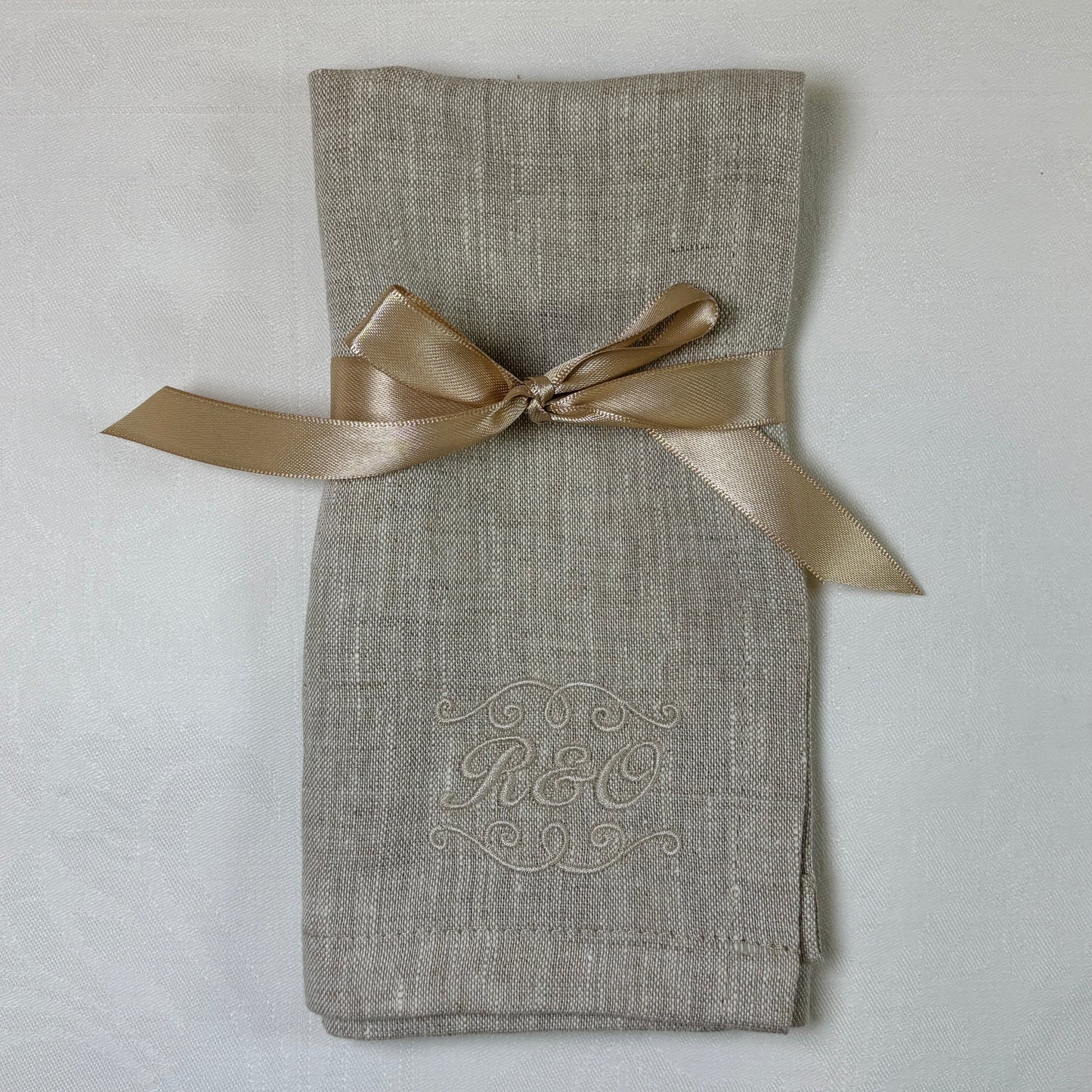 Naturfärgade melerade linneservett med ett ljusbeige band knutet med rosett. Tygservetten är broderad med beige eller naturfärgad tråd som motsvarar servettens färg.