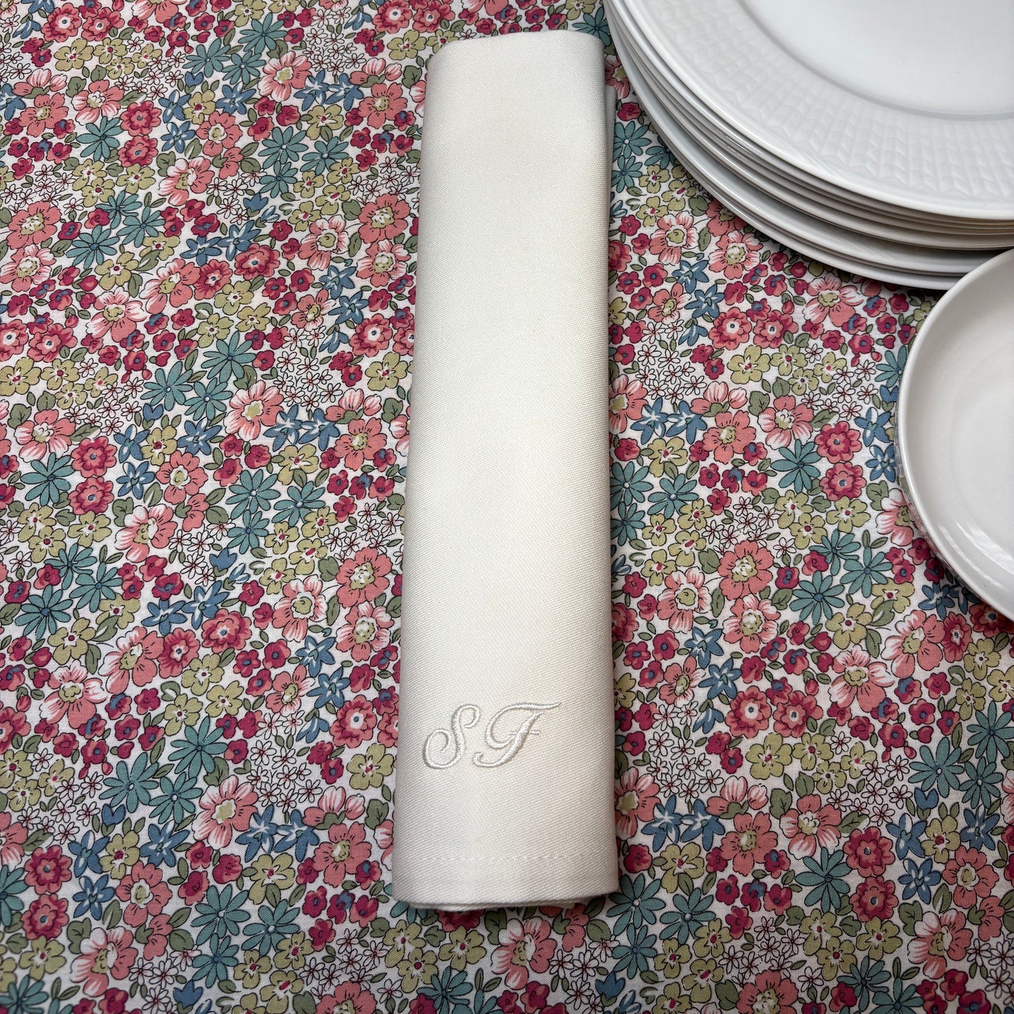 Klassiska vita tygservetter av tät och fin bomull. Initialerna SF är broderade i kursiv stil med vit blank brodyrtråd. Servetten är ihoprullad och ligger på en småblommig duk bredvid vitt porslin.