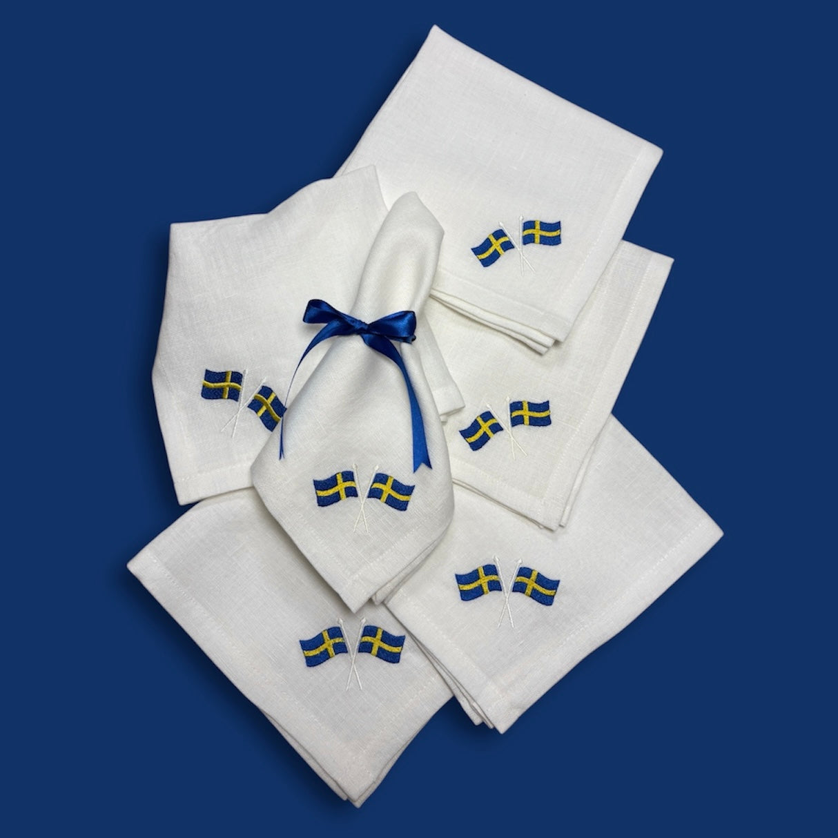 Flera vita linneservetter ligger på en blå bakgrund. Linneservetterna har två svenska flaggor broderade i hörnen. En av tygservetterna har ett blått satinband knutet med rosett.