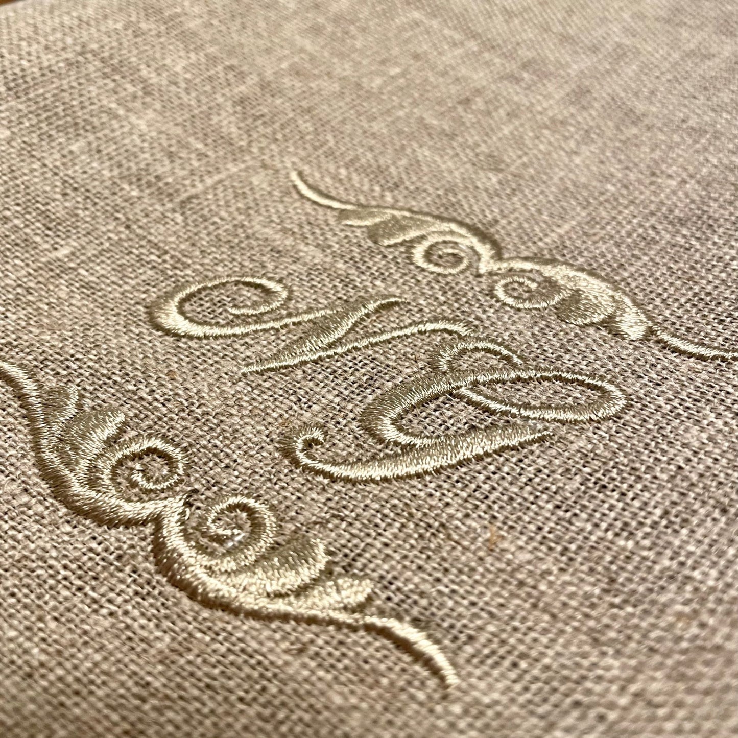 Närbild på broderat monogram på linnehanddukar. Vi broderar på beställning. Välj initialer. Varm grå-beige tråd på oblekt linne.