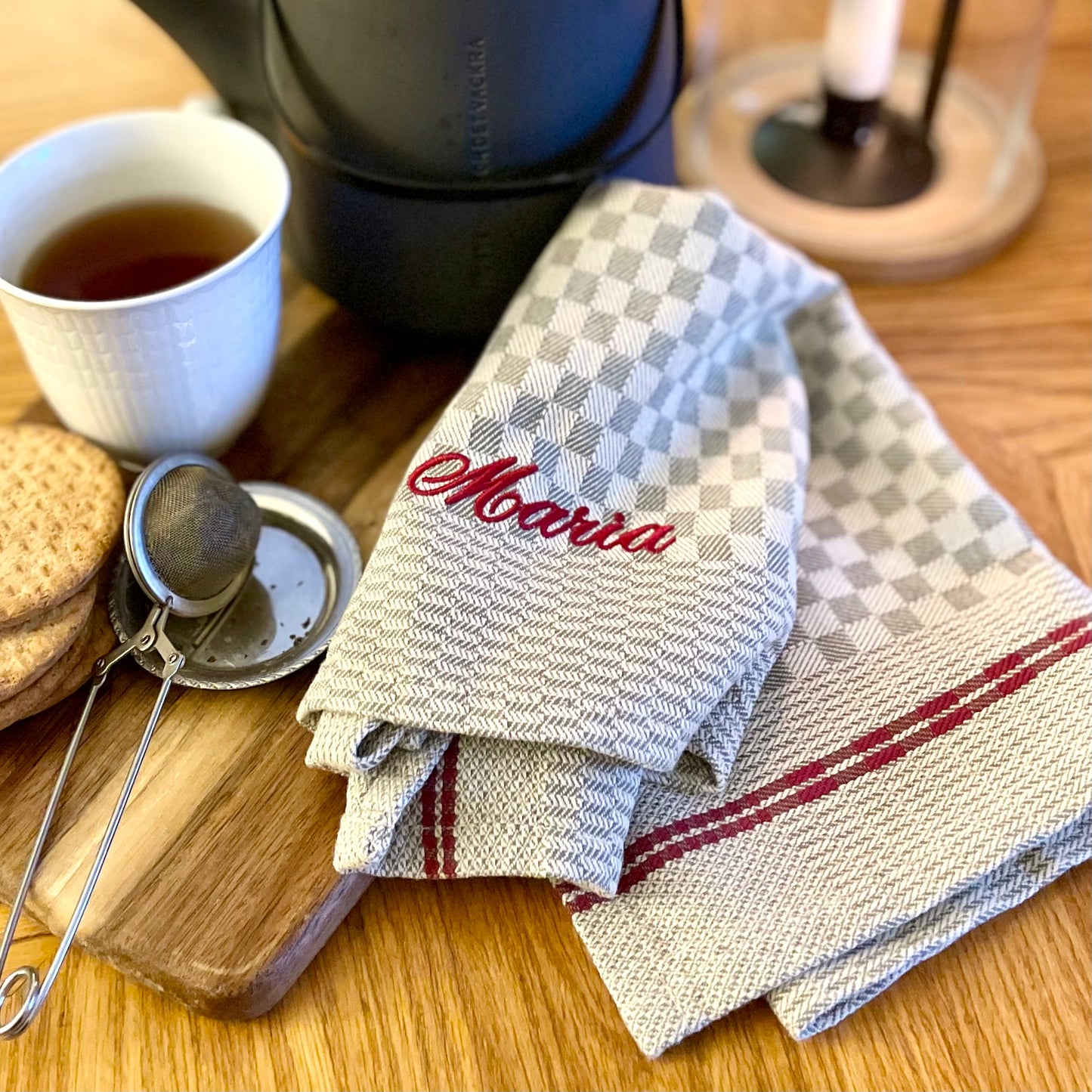 Linnefärgad rustik bakduk med vävt rutmönster och broderat namn i mörkrött. Servetten ligger på soffbord vid träbricka med tekopp och tekanna.