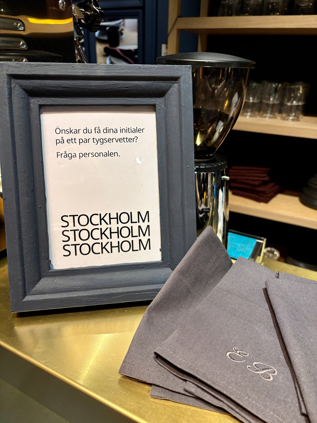 Brodering eller brodyr på event hos IKEA. Inramad informationstext om erbjudande om initialer på servetter. Grå bomullservetter med initialer i grå brodyr