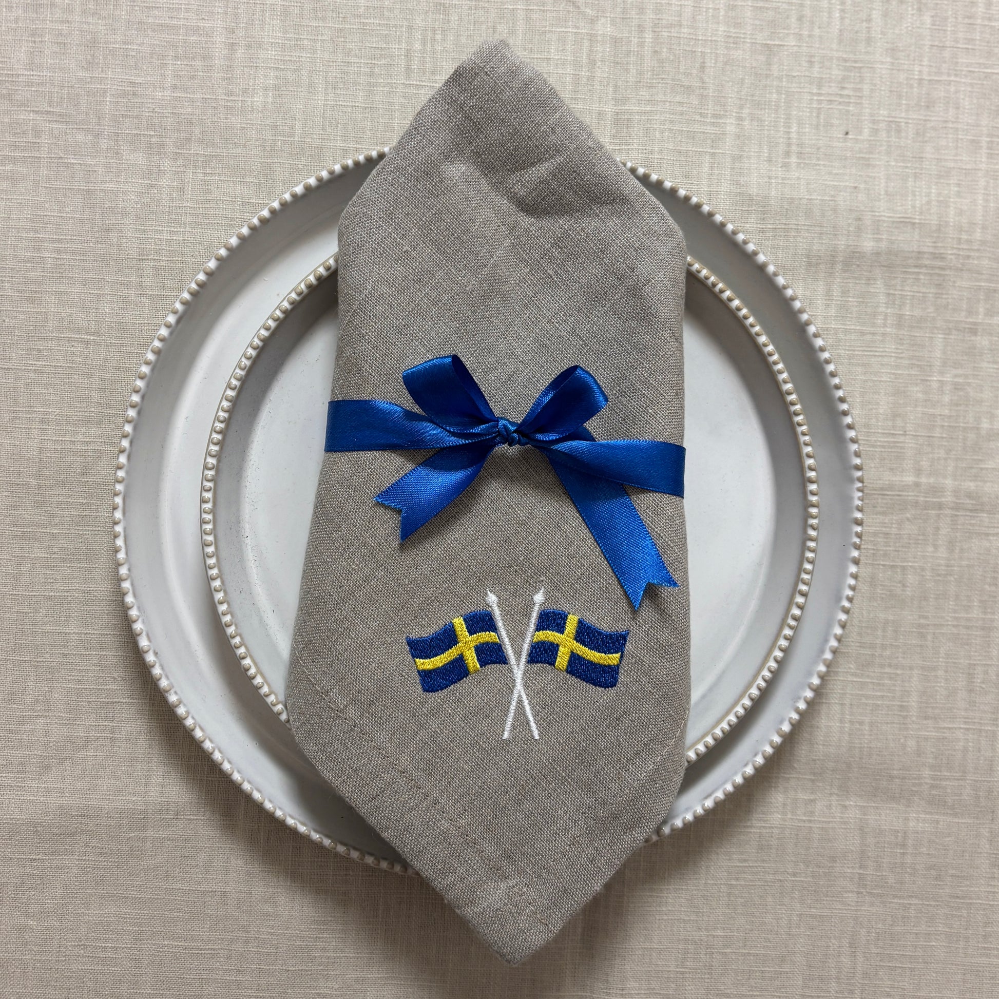En naturfärgad linneservett med två broderade svenska flaggor i hörnet är vikt och ligger på naturvita porslinstallrikar. Ett blått satinband är knutet med rosett runt linneservetten. Under allt syns en ljusbeige linneduk.