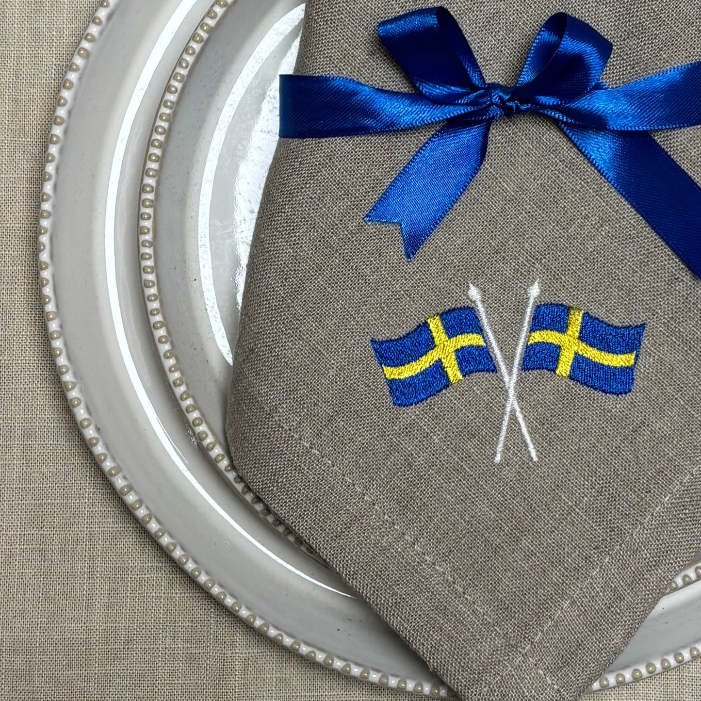 Närbild av en naturfärgad linneservett med två broderade svenska flaggor i hörnet är vikt och ligger på naturvita porslinstallrikar. Ett blått satinband är knutet med rosett runt linneservetten. Under allt syns en ljusbeige linneduk.