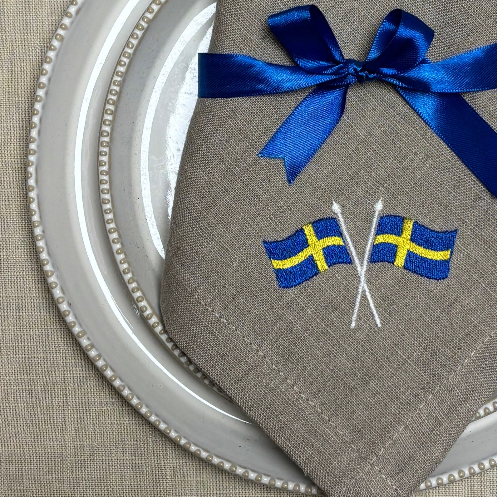 Närbild av en naturfärgad linneservett med två broderade svenska flaggor i hörnet är vikt och ligger på naturvita porslinstallrikar. Ett blått satinband är knutet med rosett runt linneservetten. Under allt syns en ljusbeige linneduk.
