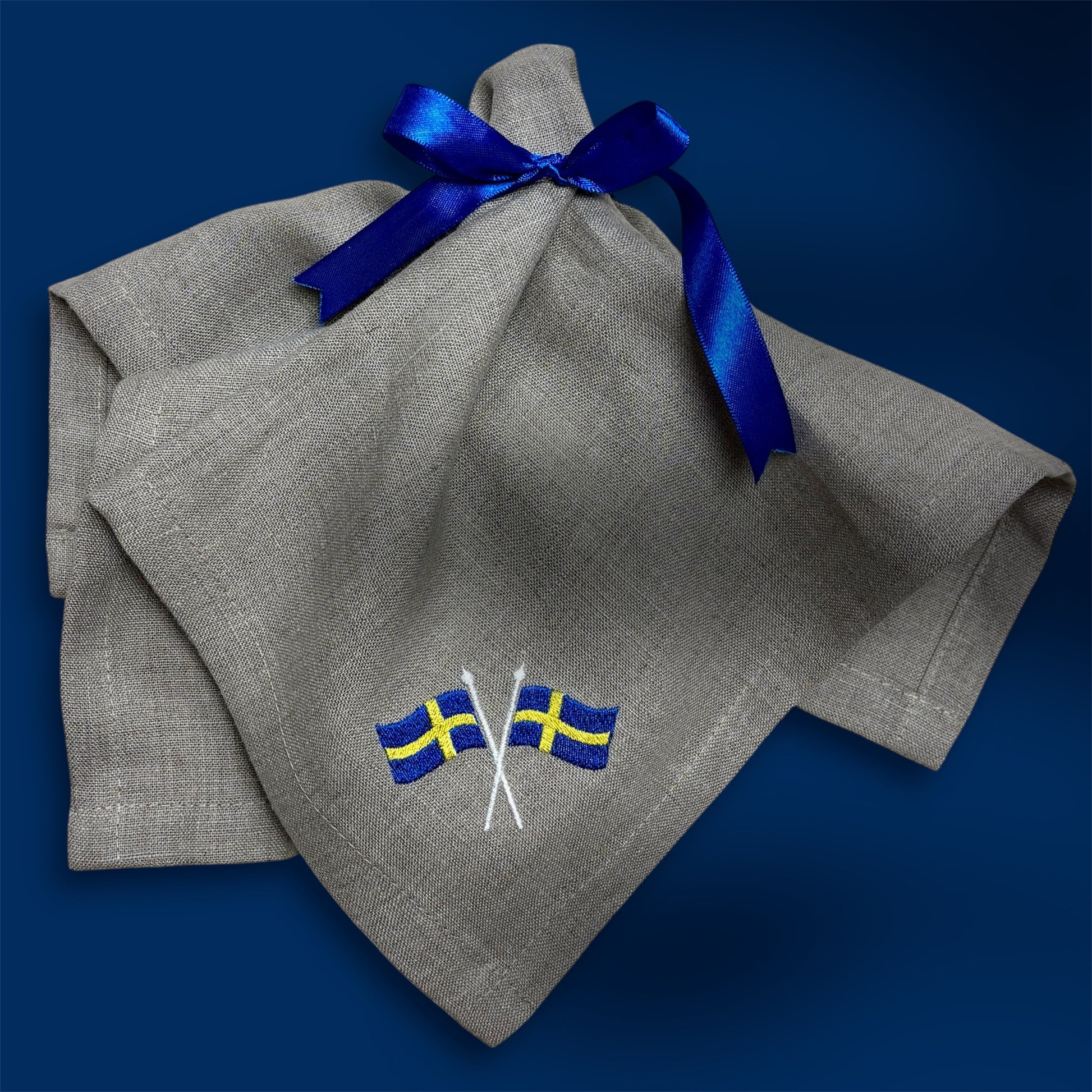 En naturfärgad linneservett med två broderade svenska flaggor i hörnet är vikt och ligger på en blå bakgrund. Ett blått satinband är knutet med rosett runt linneservetten. 