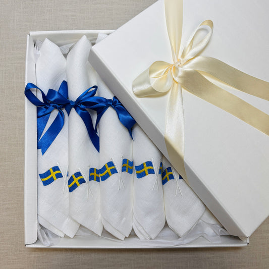 Krämvit presentask är öppen och visar 6 vita linneservetter. Linneservetterna är broderade med svenska flaggor. Varje servett har ett blått satinband knutet i rosett. Den krämvita asken har ett krämvitt satinband knutet runt locket.