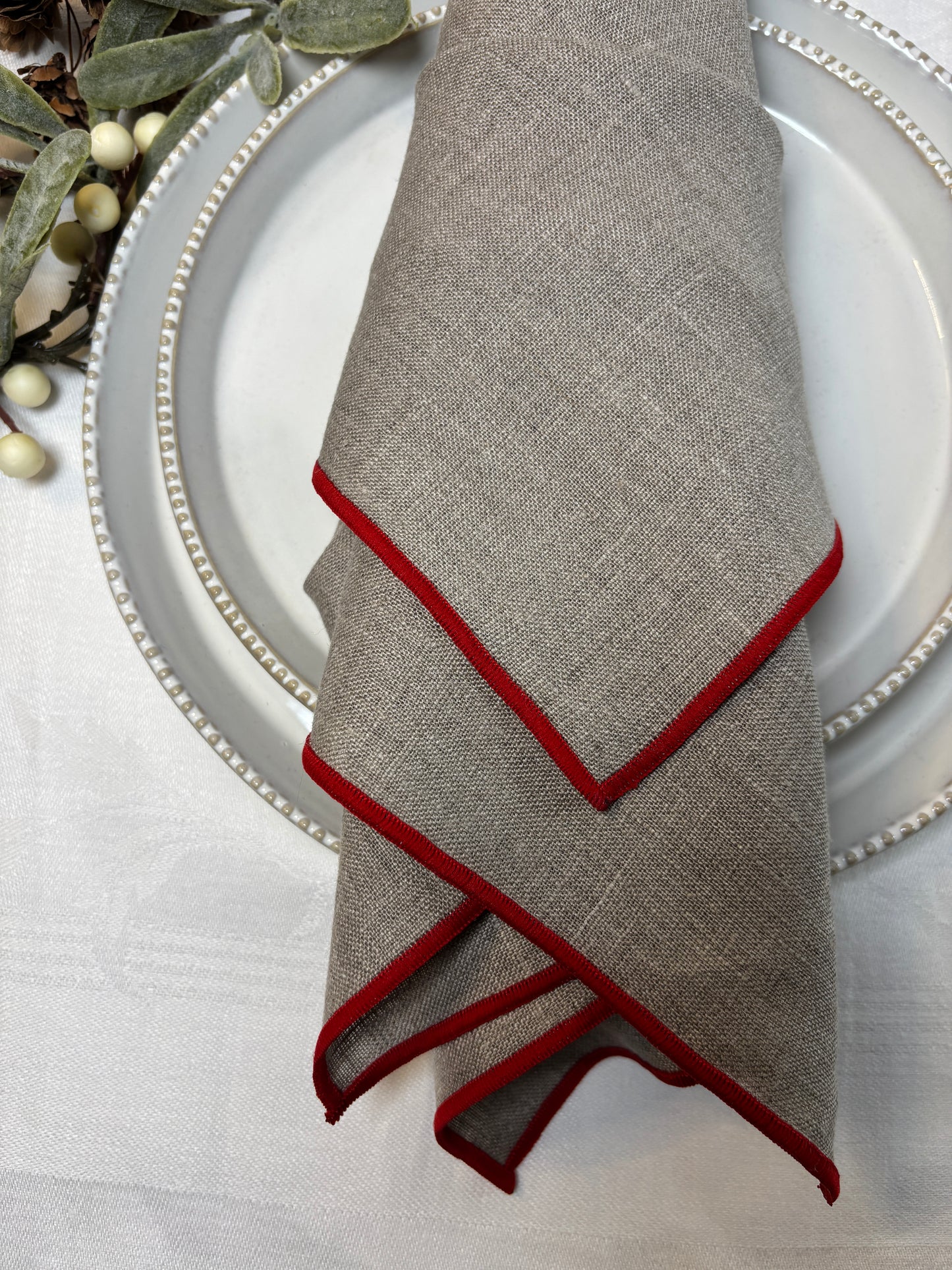 Naturfärgad linneservett med röda smala kanter. Ligger vikt mitt på tallrik och assiett på vit linneduk.