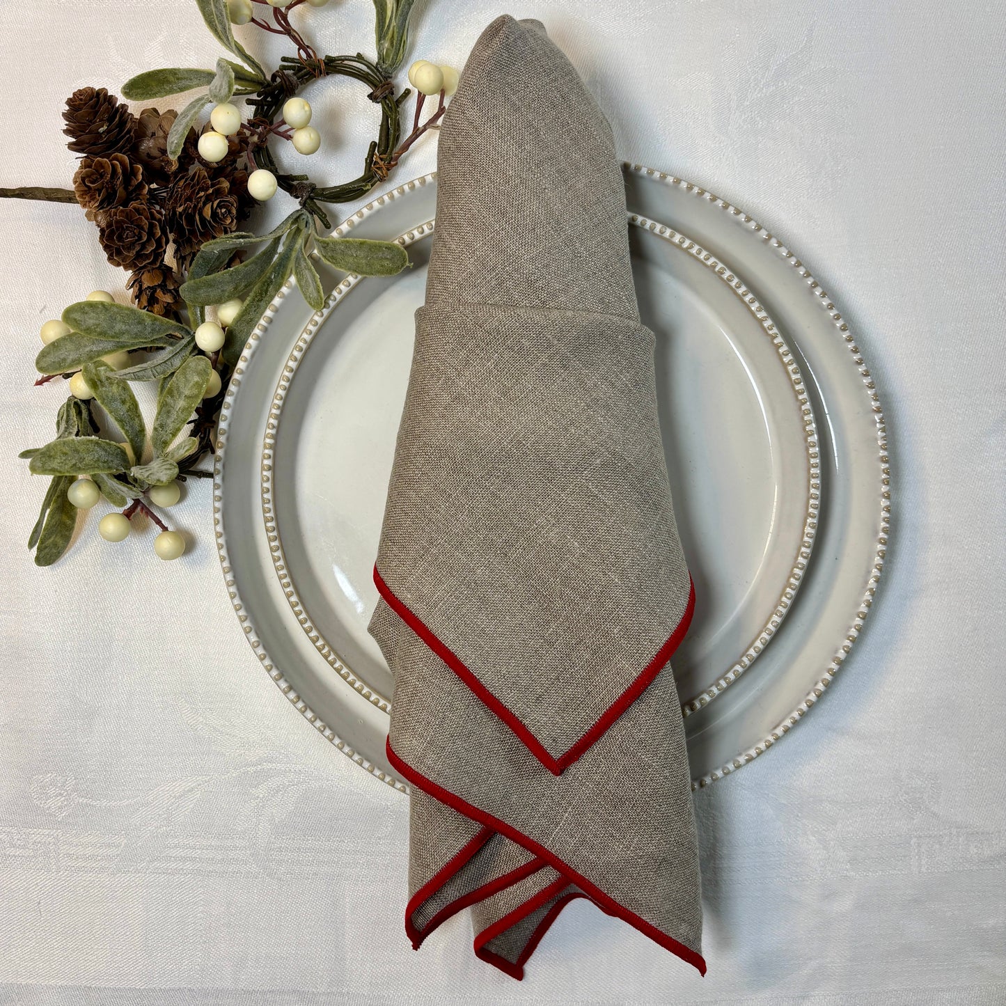 Naturfärgad linneservett med röda smala kanter. Ligger vikt mitt på tallrik och assiett på vit linneduk. 