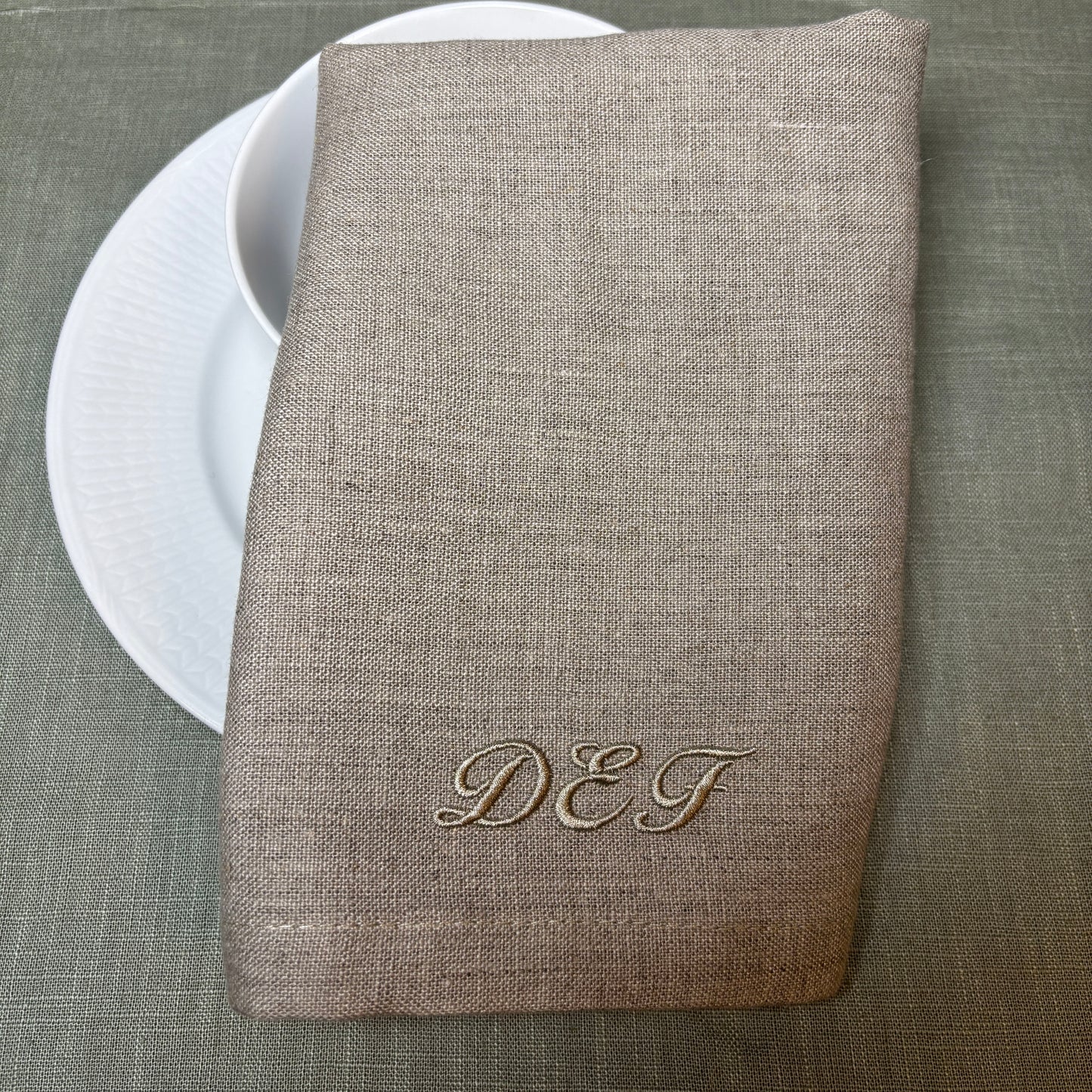Tygservetter av linne med broderat monogram eller initialer i samma nyans som linneservetten. Naturvärgad eller linnefärgad. Initialerna DEF. Bakom syns vitt porslin och en grön duk.
