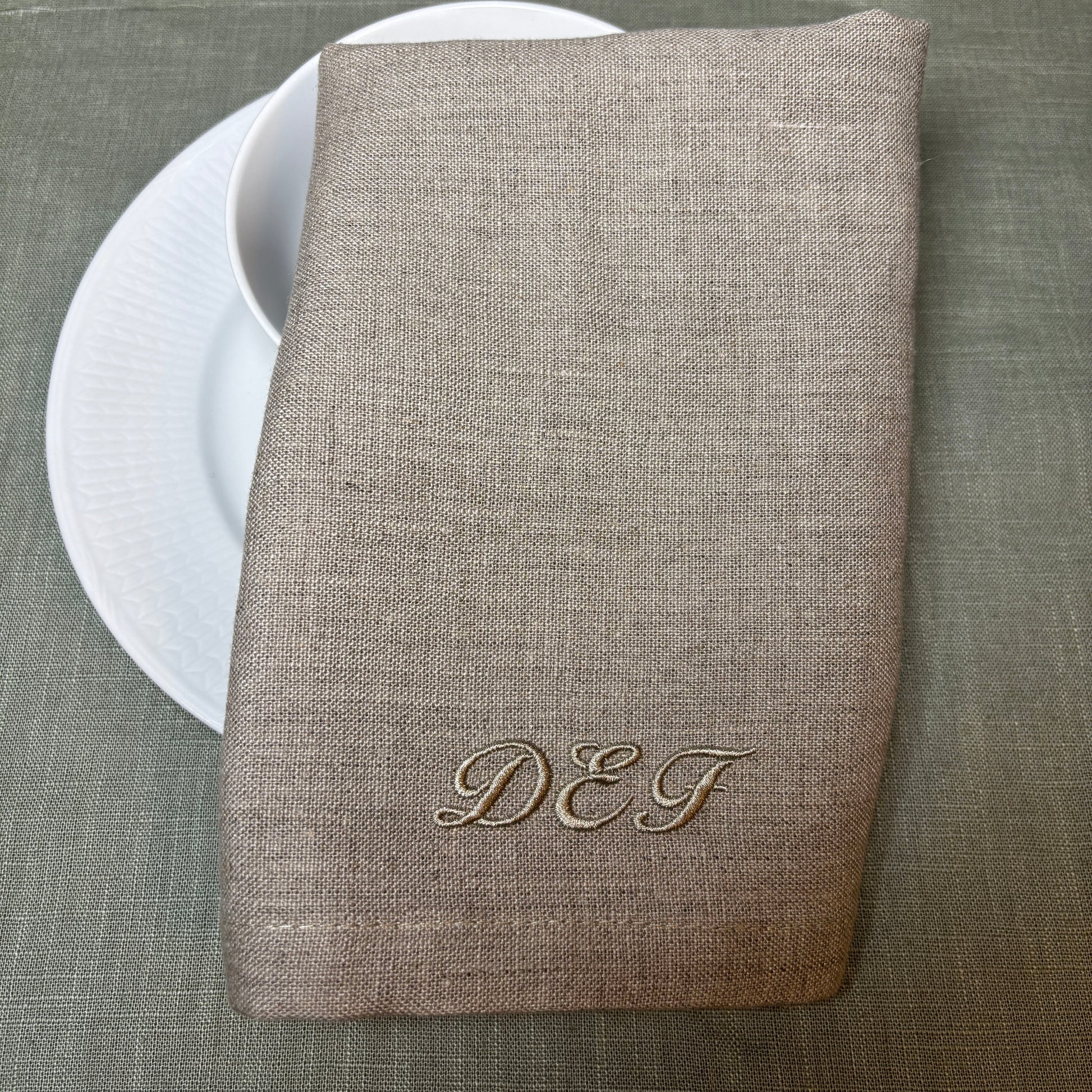 Tygservetter av linne med broderat monogram eller initialer i samma nyans som linneservetten. Naturvärgad eller linnefärgad. Initialerna DEF. Bakom syns vitt porslin och en grön duk.