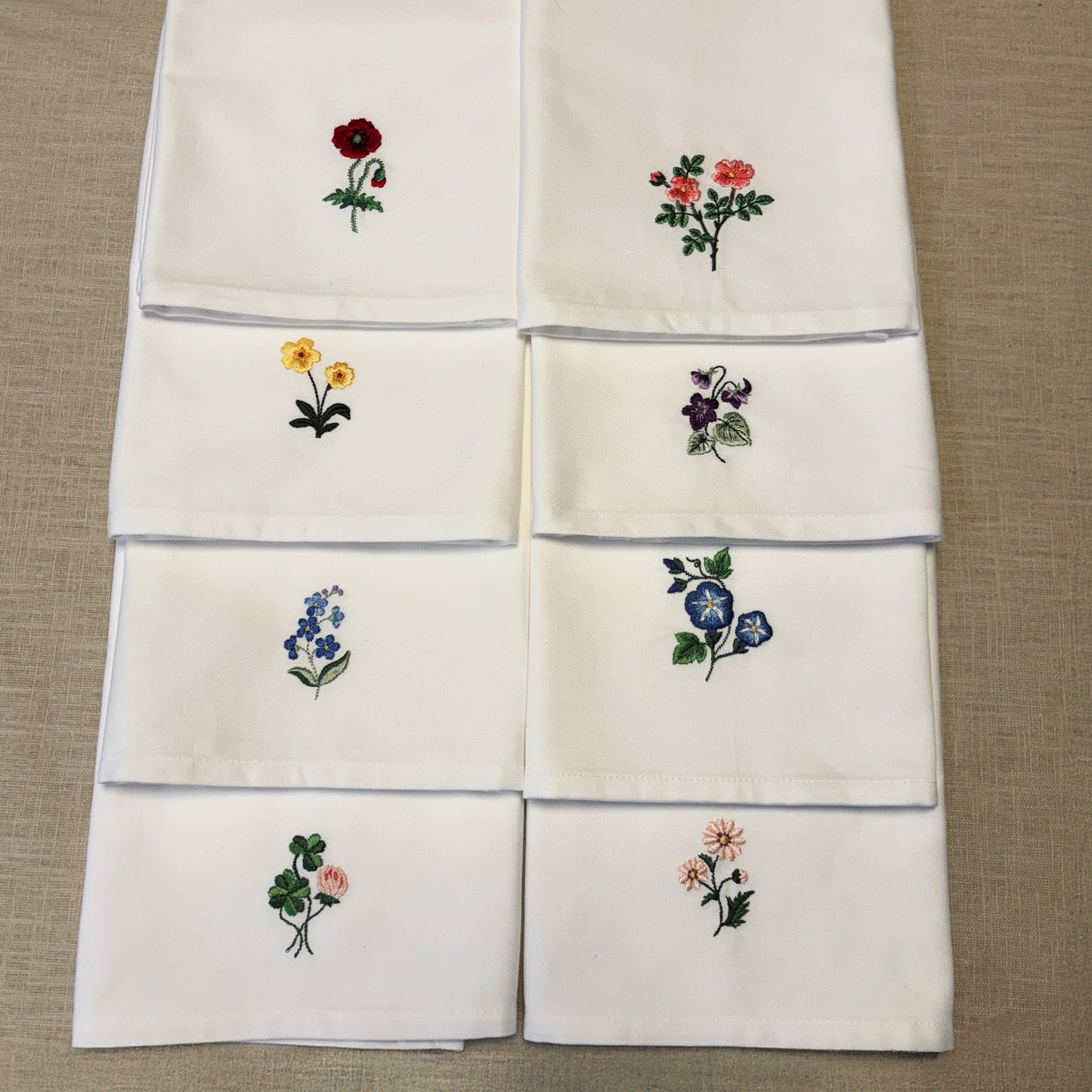 8 stycken vita tygservetter med olika blommor broderade på vardera. Ligger på en beige linneduk. Servetternas blommor är Klöver, forgetmenot, Primrose, pion, tusensköna, vildros, MorningGlory och Borage