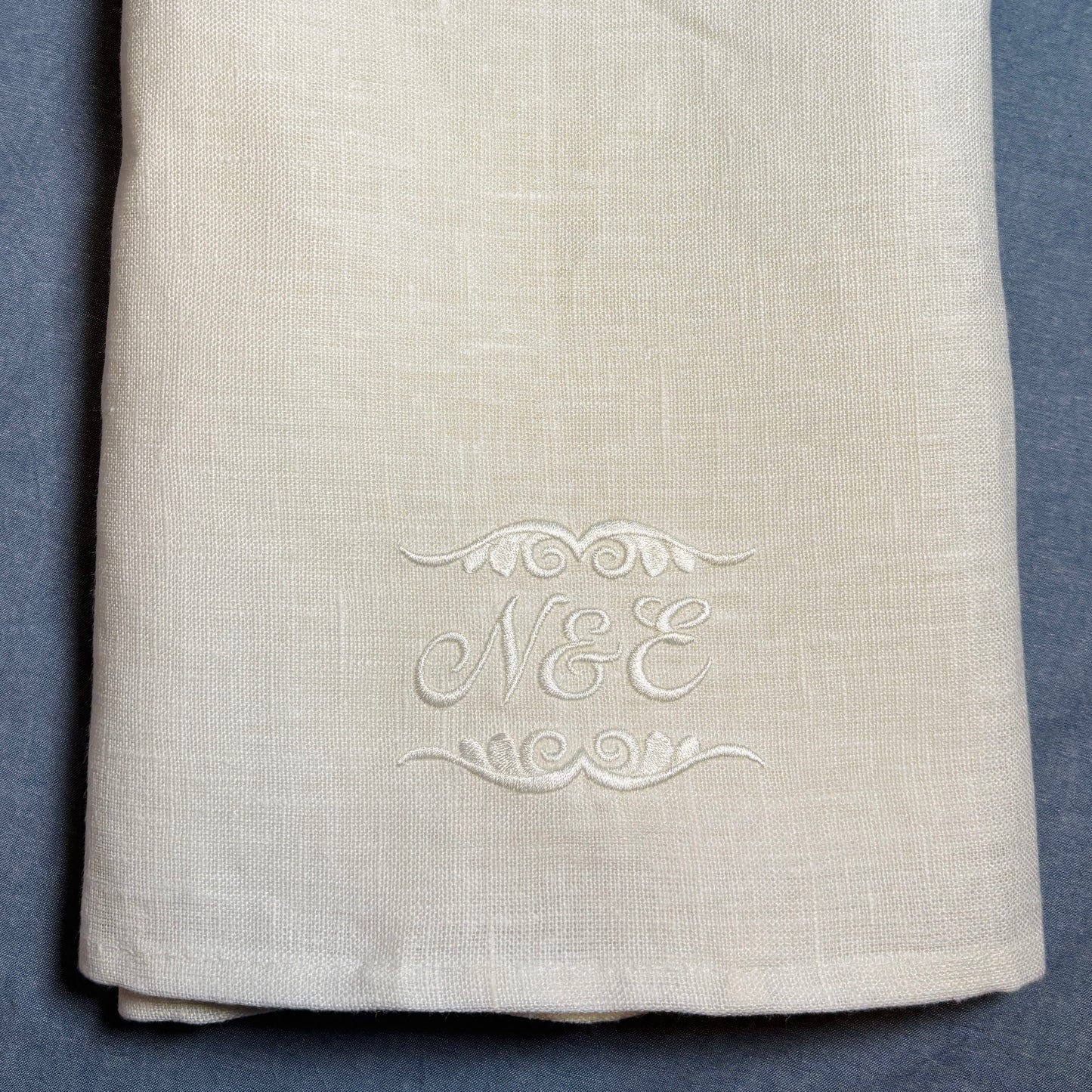 Närbild av en vit (off white) linnehandduk med broderade initialer och dekor av vit blank brodyrtråd nederst. Närbild för att visa brodyren som är kursiv och snirklig stil N&E samt dekor ovanför och under bokstäverna. Linnehandduken ligger på en blå duk.