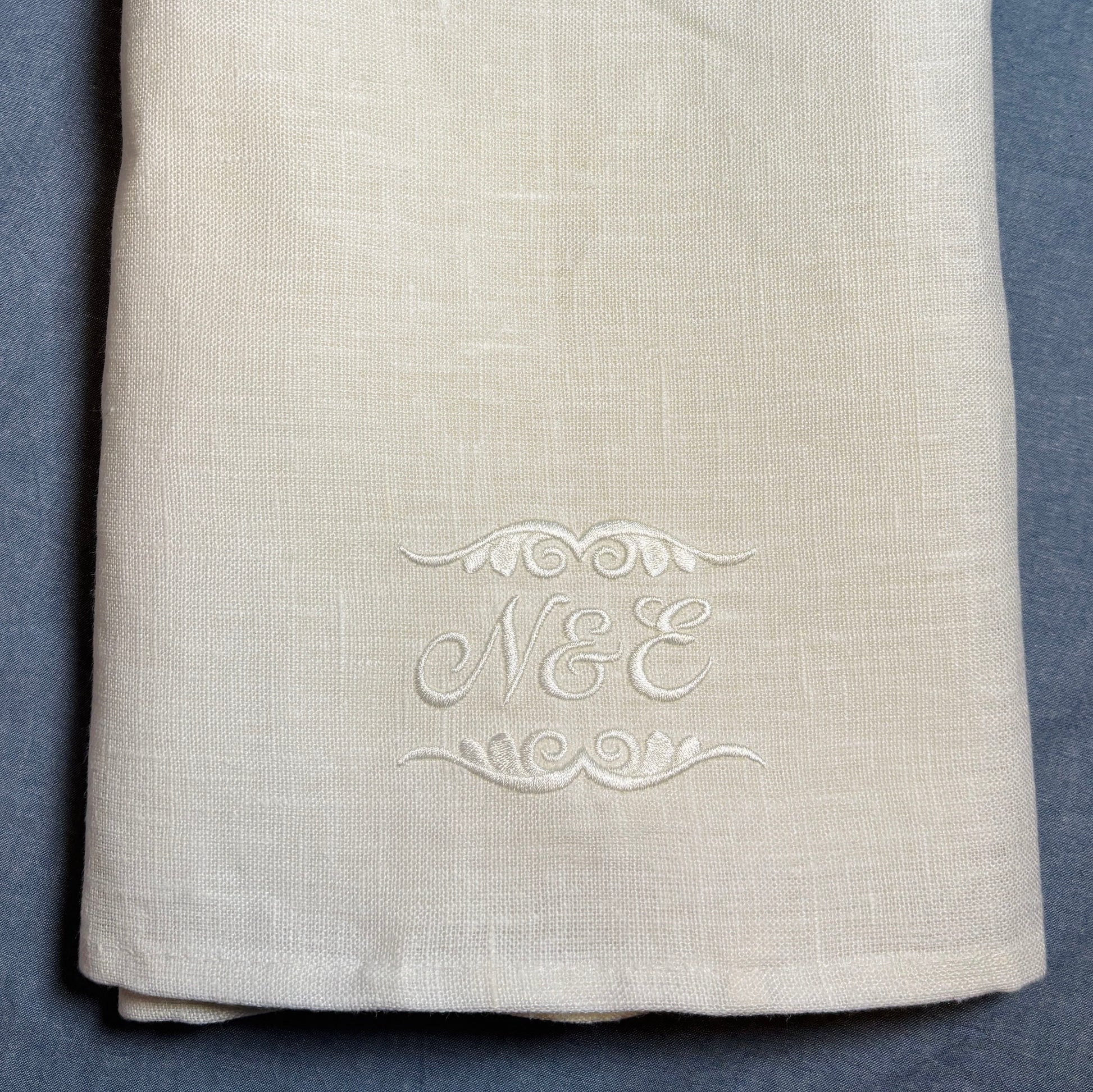 Närbild av en vit (off white) linnehandduk med broderade initialer och dekor av vit blank brodyrtråd nederst. Närbild för att visa brodyren som är kursiv och snirklig stil N&E samt dekor ovanför och under bokstäverna. Linnehandduken ligger på en blå duk.