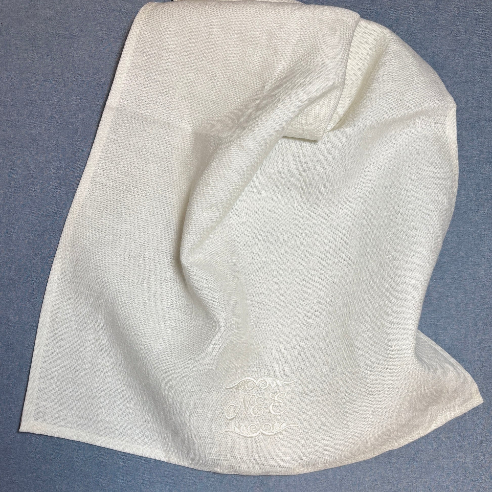 Vit (off white) linnehandduk med broderade initialer och dekor av vit blank brodyrtråd nederst. N&E. Dekoren är ovanför och under N&E. Linnehandduken ligger på en blå duk.