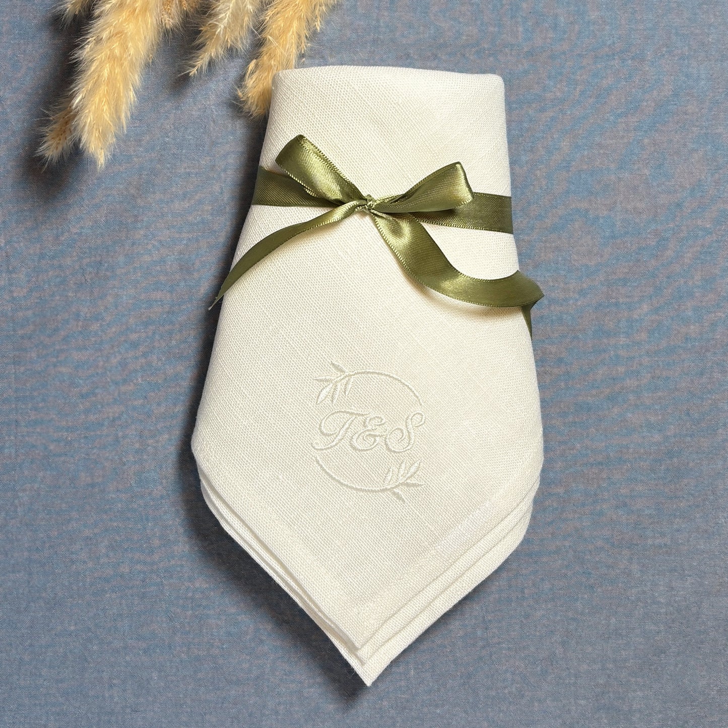 Vit tygservett av 100% linne med vit brodyr i hörnet. Bokstäverna T&S i cirkel av olivkvistar. Ligger vikt på en blå duk. Den vita linneservetten har en ljus olivgrön satinband knutet med rosett.