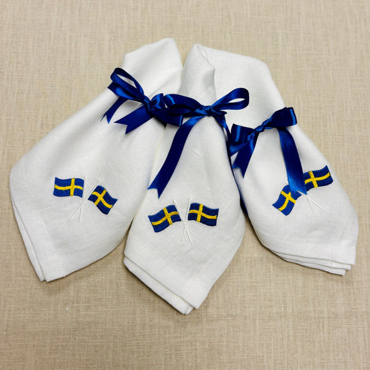 Tre vita linneservetter som är broderade med svenska flaggor ligger på en ljusbeige linneduk. Alla tre tygservetterna har blå satinband knutna i rosetter. 