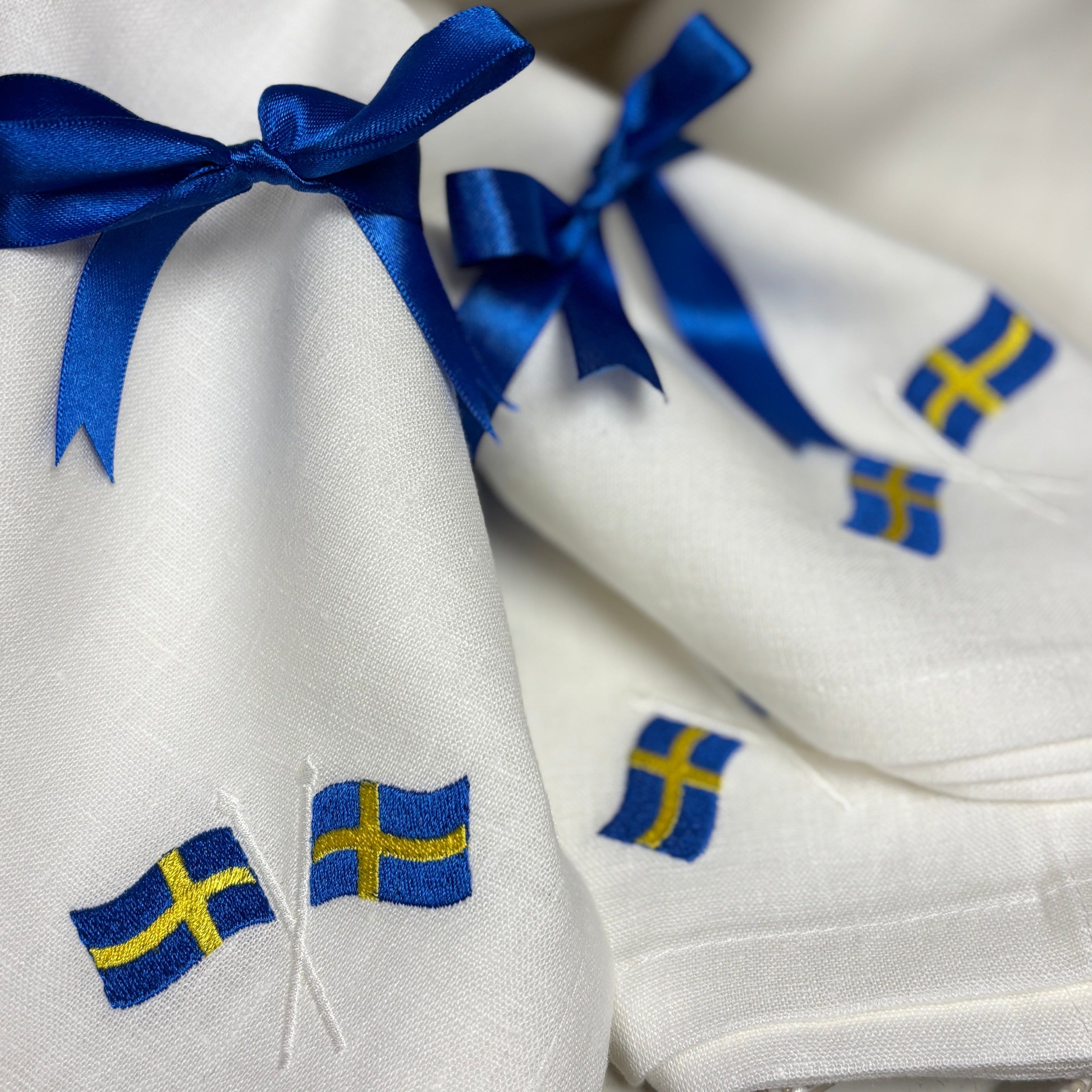 Närbild av vita linneservetter som har broderade svenska flaggor. Blåa satinband är knutna runt servetterna .