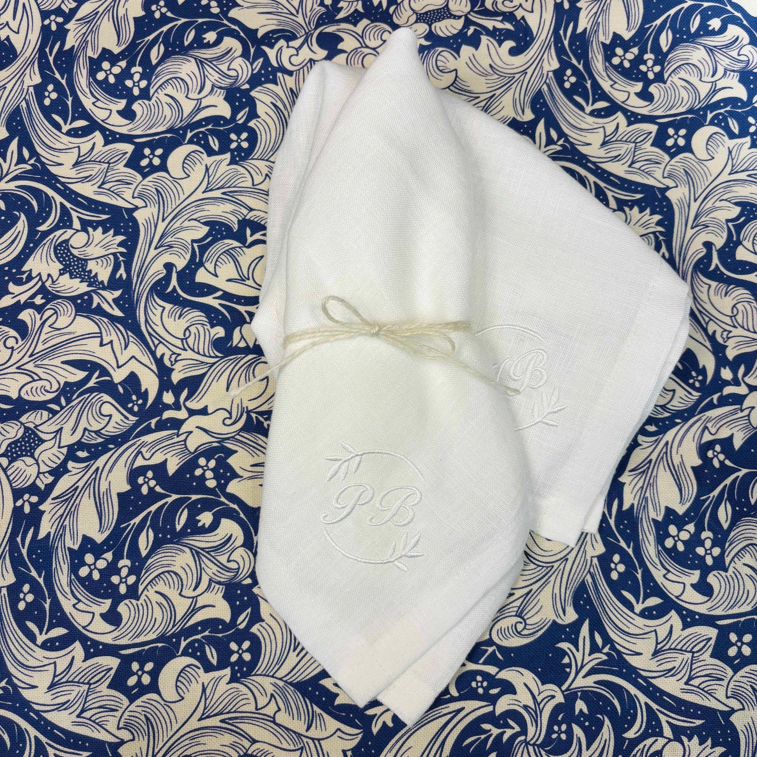 Vit tygservett av 100% linne linneservetter med vit brodyr i hörnet. Bokstäverna PB i cirkel av olivkvistar. Ligger på en William Morris mönstrad duk som är blå och vit . 