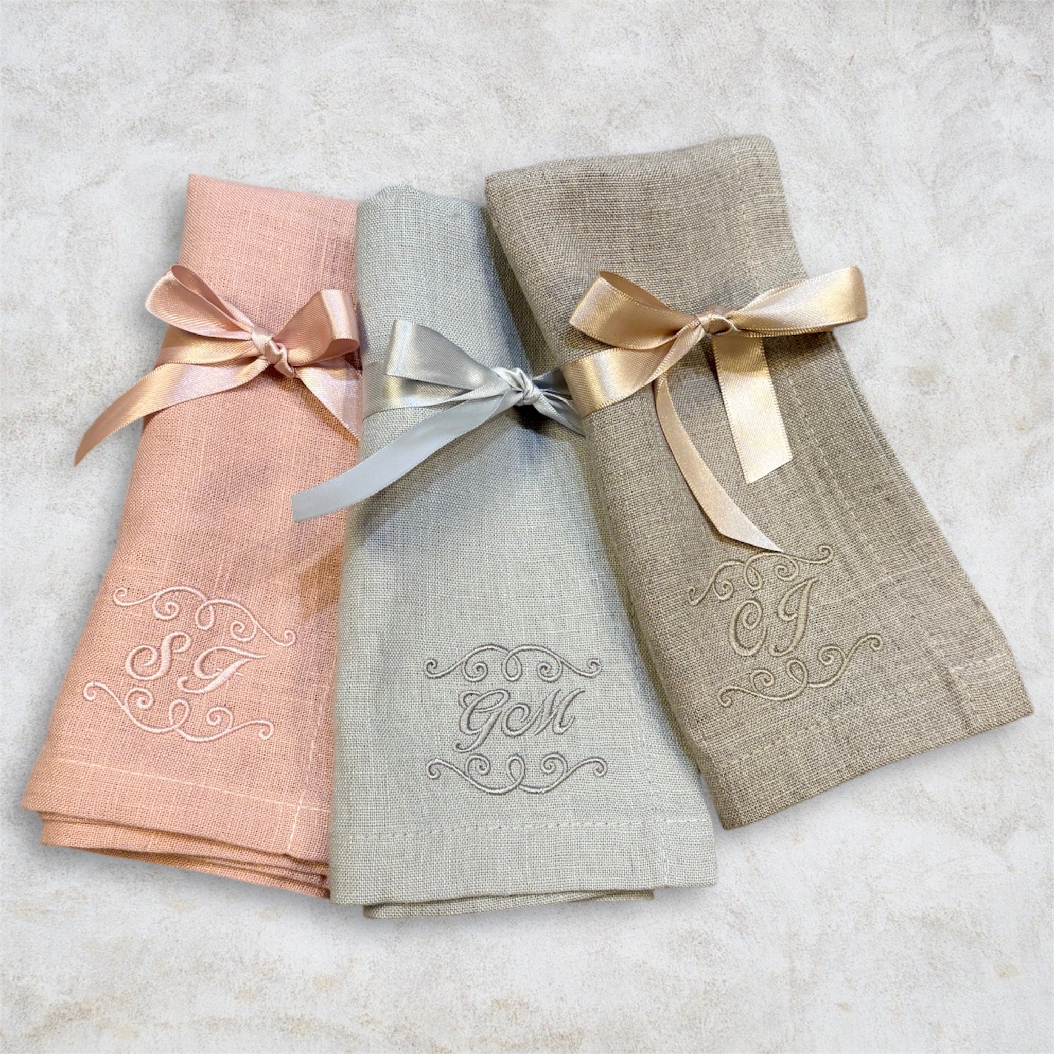 Tre linneservetter med olika initialer broderade. Rosa, ljusgrå och naturfärgade. Alla tre tygservetter har sidenband runt som är knutna till rosetter. Servetterna är vikta så de broderade monogrammen visas upp.