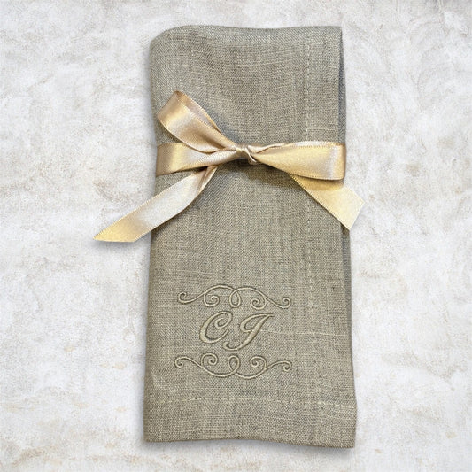 Naturfärgade linneservett med ett ljusbeige band knutet med rosett. Tygservetten är broderad med beige eller naturfärgad tråd som motsvarar servettens färg. Initialer samt dekor. Linneservetten är vikt så den broderade kanten med initialerna CJ visas upp.