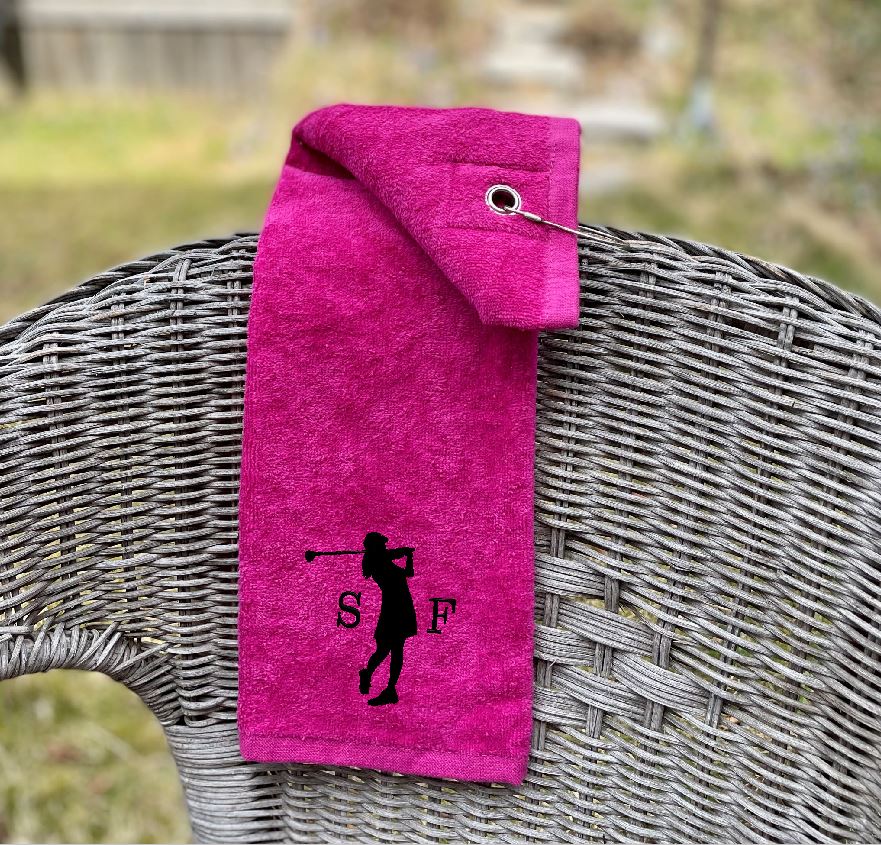 Cerise rosa golfhandduk med initialer broderat samt dam som spelar golf. Golfsving. Svart brodyr på cerise golfhandduk.